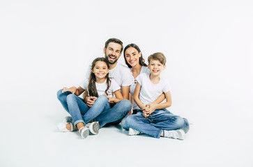 Relationship concept. Beautiful and happy smiling young family in white T-shirts are hugging and have a fun time together while sitting on the floor and looking on camera.