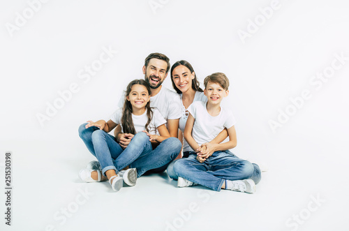 Relationship concept. Beautiful and happy smiling young family in white T-shirts are hugging and have a fun time together while sitting on the floor and looking on camera.