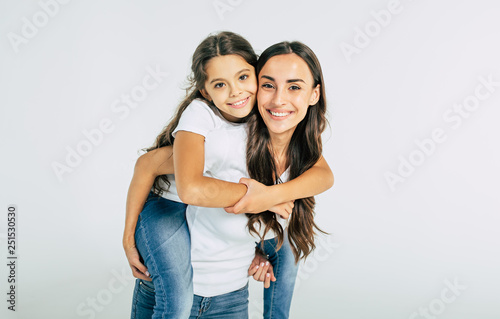 Beautiful young brunette mother holds on back her cute little daughter in white T-shirts isolated in studio