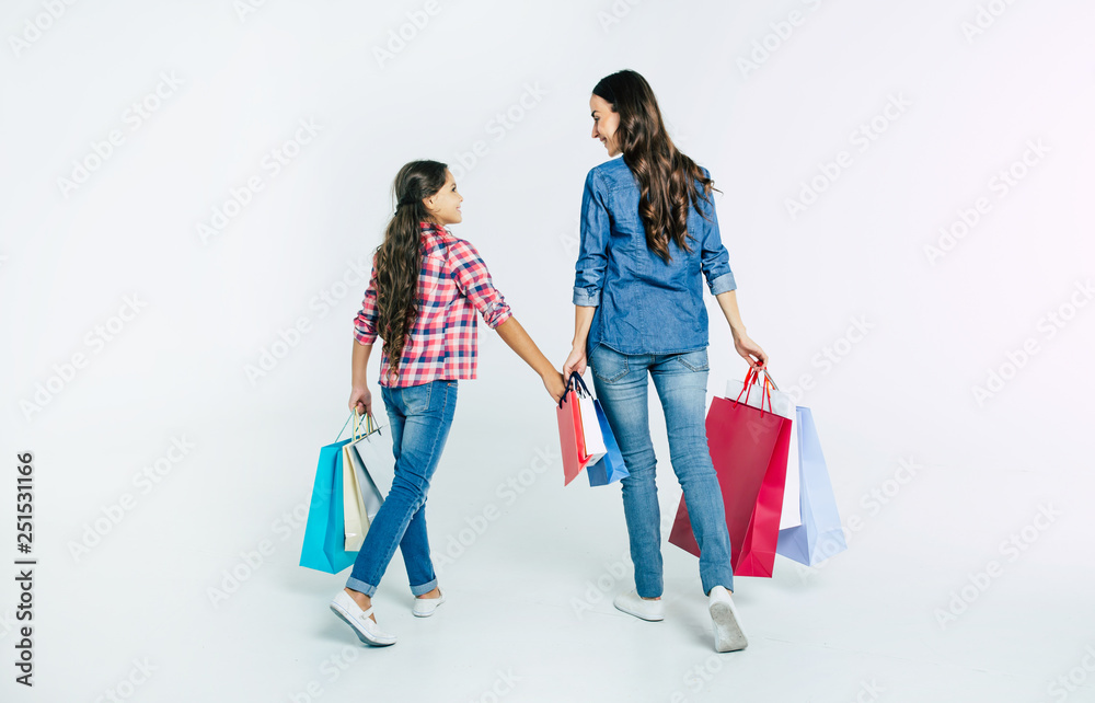 Beautiful mother and cute daughter in casual clothes with shopping bags in hands make purchases at the mall isolated on white Beautiful mother and cute daughter in casual clothes with shopping bags in hands make purchases at the mall isolated on white