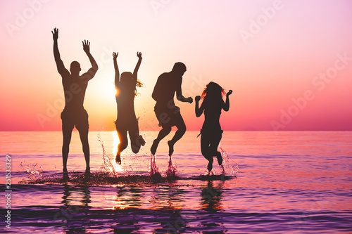 Happy friends jumping inside water on tropical beach at sunset