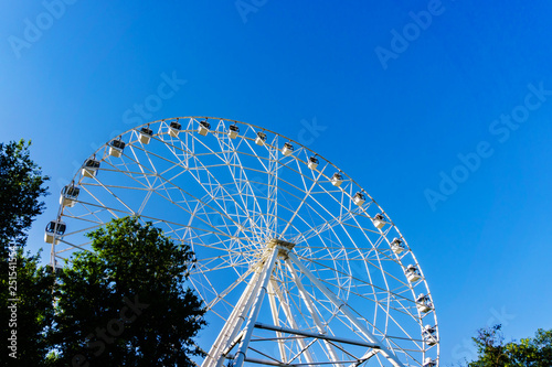 A new very tall white Ferris wheel with comfortable booths for people is slowly rotating in a circle in the center of a big city against a clear blue sky