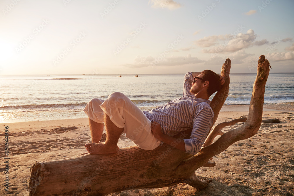 Modern man posing on the wooden stump on the sandy beach.