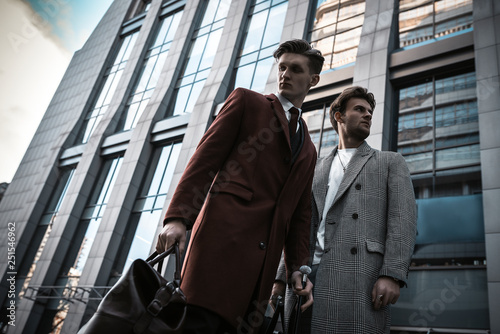 Two fashion men models posing on city street. Wearing in classic and casual closes. Suit, coat, shirt, sweater, boots and leather bag. Men model test near business center