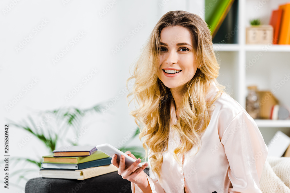 Laughing blonde woman in pyjamas holding smartphone and looking at camera Laughing blonde woman in pyjamas holding smartphone and looking at camera