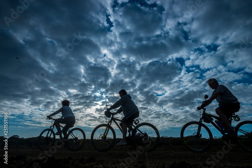 Group of people riding mountain bike during a beautiful dark cloudy sunset - outdoor nature sport activity together for healhty lifestyle - three bikers in silhouette - cold filter