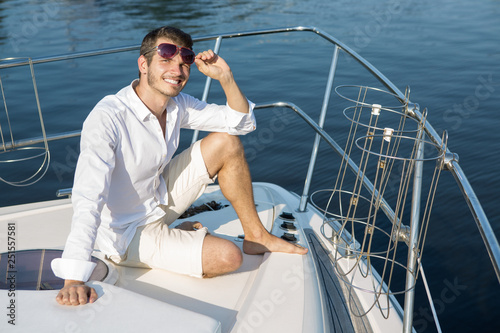 Bearded man with charming smile sitting on boat bow and posing. Male model wearing white shirt and shorts and black sunglasses. He looking at camera, smiling and holding sunglasses with one hand.