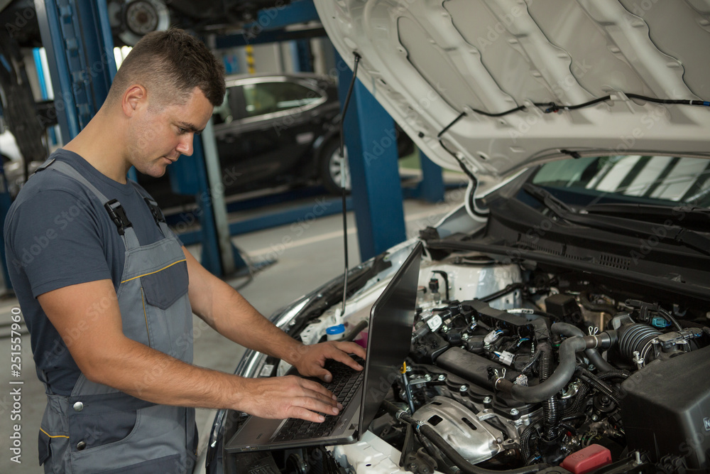 Side view of skilled repairman typing in computer and checking serviceability of automobile at car service. Thoughtful male in grey uniform standing near white car with open hood and looking at pc. Side view of skilled repairman typing in computer and checking serviceability of automobile at car service. Thoughtful male in grey uniform standing near white car with open hood and looking at pc.
