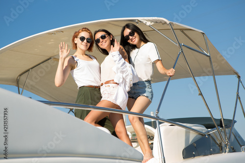 Three beautiful and slender girls posing and standing on yacht. Charming women smiling and looking at camera. Two brunettes and one woman with ginger hair wearing white clothes and sunglasses.