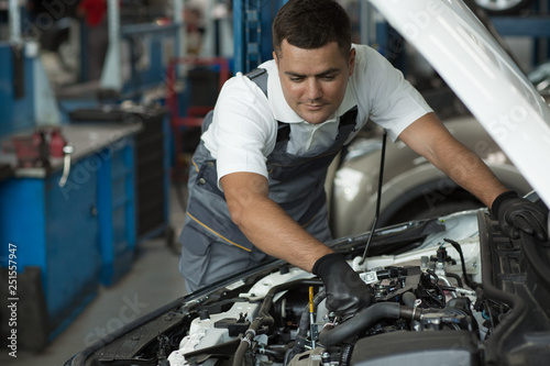 Professional mechanic leaning over open hood of car and fixing breakage at auto station. Man in white shirt and grey uniform in process of checking serviceability of vehicle. Concept of car repair.