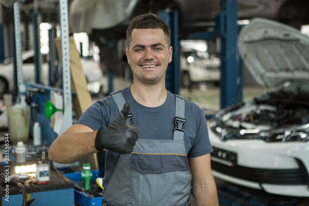 Professional male worker in uniform and protective gloves standing near car, showing sign okay, looking at camera and smiling. Positive mechanic enjoying his job at auto service. Concept of happiness.