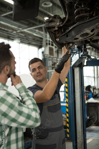 Skilled mechanic explaining male client problem with car in automobile service. Repairman in uniform and gloves pointing with hand at lifted vehicle and talking with thoughtful man standing near.