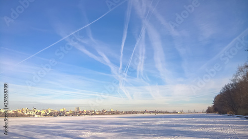 Traces of the aircraft in a clear, blue sky over the frozen river against the background of the city.