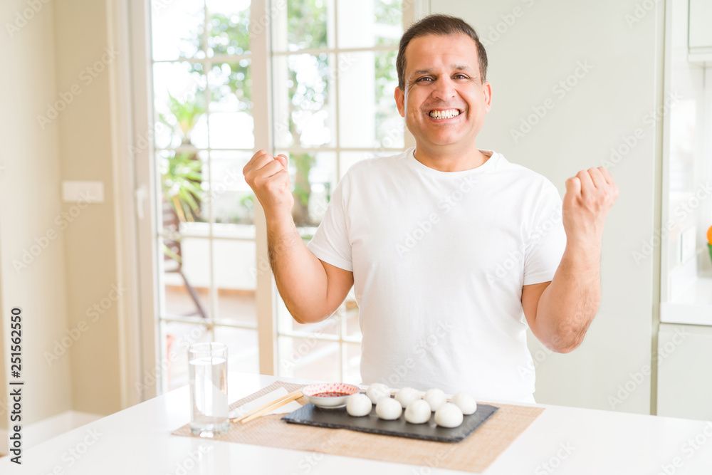 Middle age man eating asian dumplings at home celebrating surprised and amazed for success with arms raised and open eyes. Winner concept. Middle age man eating asian dumplings at home celebrating surprised and amazed for success with arms raised and open eyes. Winner concept.