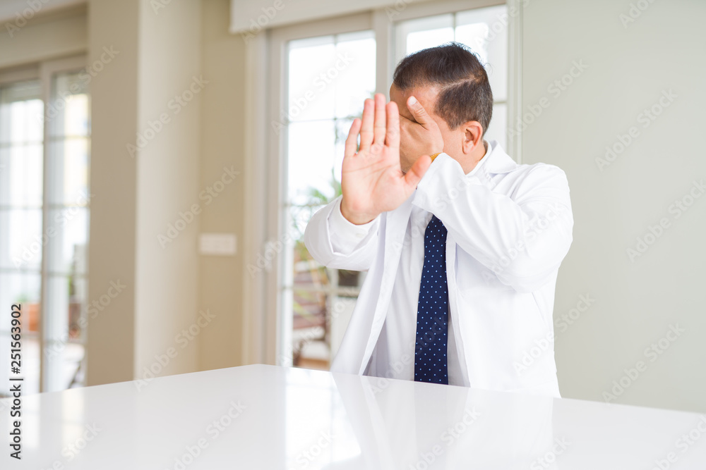 Middle age doctor man wearing medical coat at the clinic covering eyes with hands and doing stop gesture with sad and fear expression. Embarrassed and negative concept.