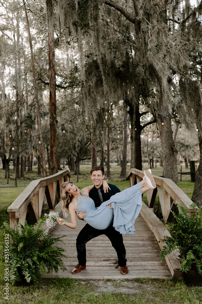 Fototapeta premium Beautiful Bridesmaid Woman in Blue Dress and Bouquet with Her Date at a Formal Wedding Party Celebration Event Outside in the Woods Taking Couple Portraits on Small Wooden Bridge