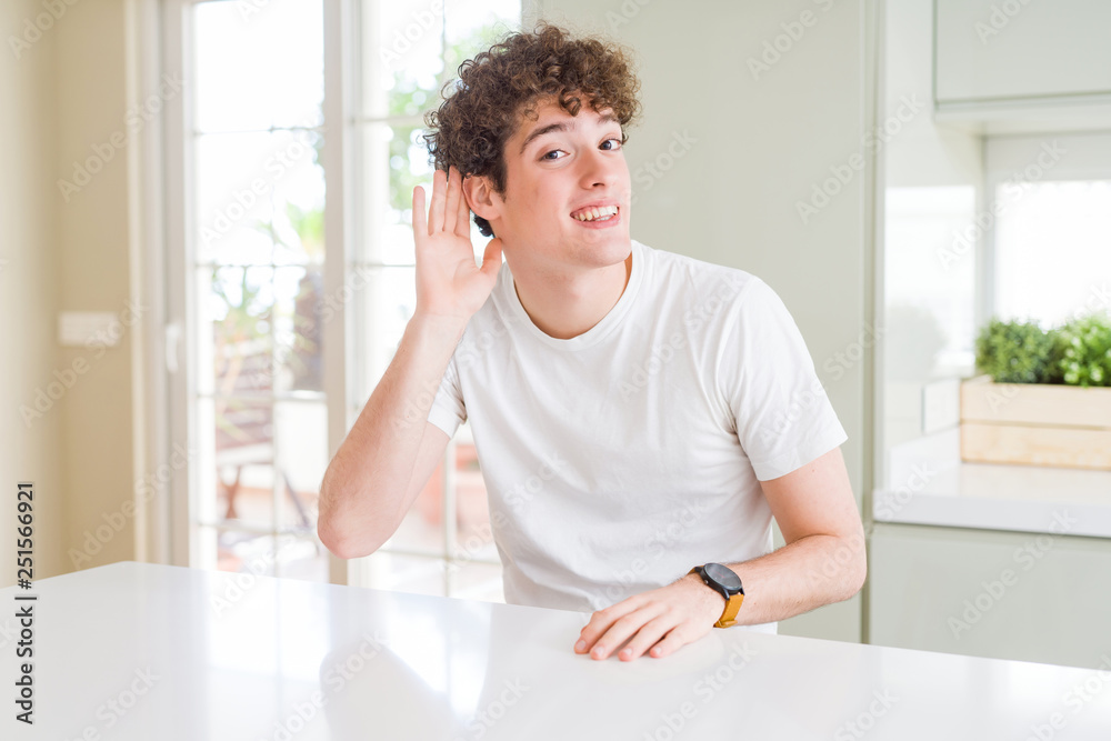 Young handsome man wearing white t-shirt at home smiling with hand over ear listening an hearing to rumor or gossip. Deafness concept. Young handsome man wearing white t-shirt at home smiling with hand over ear listening an hearing to rumor or gossip. Deafness concept.