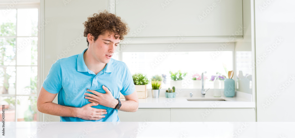 Wide shot of young handsome man at home with hand on stomach because indigestion, painful illness feeling unwell. Ache concept. Wide shot of young handsome man at home with hand on stomach because indigestion, painful illness feeling unwell. Ache concept.