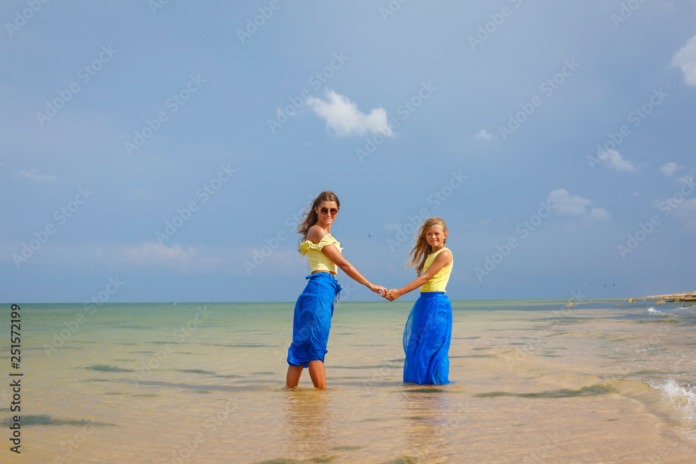 Pretty young mother and her cheerful daughter with long hair in blue skirt walking in ocean near the shore during their vacation