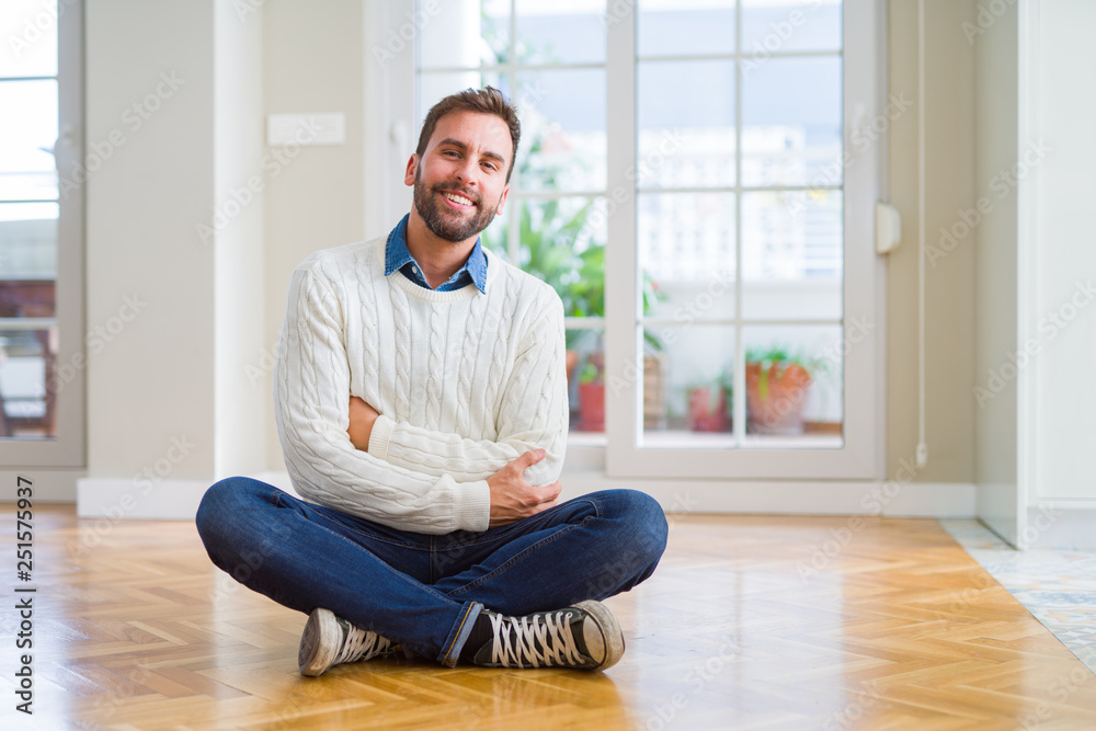 Handsome man wearing casual sweater sitting on the floor at home happy face smiling with crossed arms looking at the camera. Positive person.