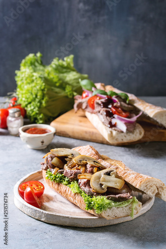 Beef baguette sandwich with champignon mushrooms, green salad, fried onion served on ceramic plate with ingredients above over grey blue table.