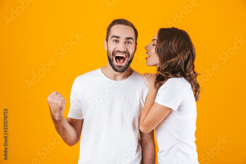Image of brunette woman whispering secret or interesting gossip to man in his ear, isolated over yellow background