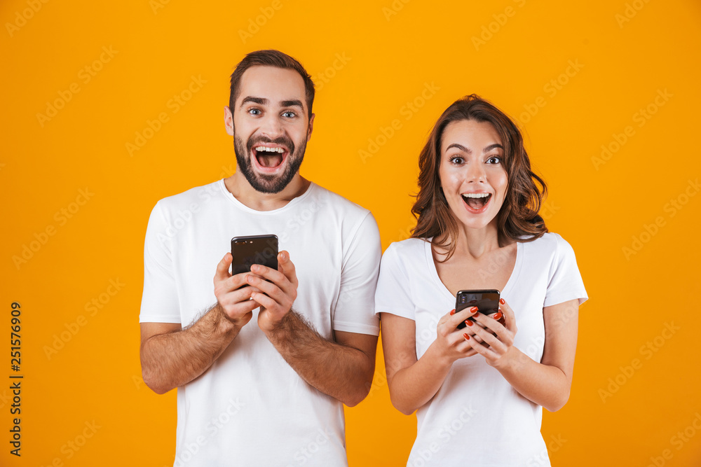 Image of beautiful couple man and woman smiling while both using mobile phones, isolated over yellow background Image of beautiful couple man and woman smiling while both using mobile phones, isolated over yellow background