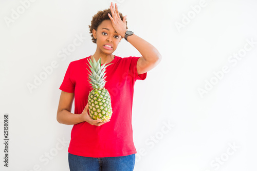 Young african american woman holding fresh healthy pineapple fruit stressed with hand on head, shocked with shame and surprise face, angry and frustrated. Fear and upset for mistake.