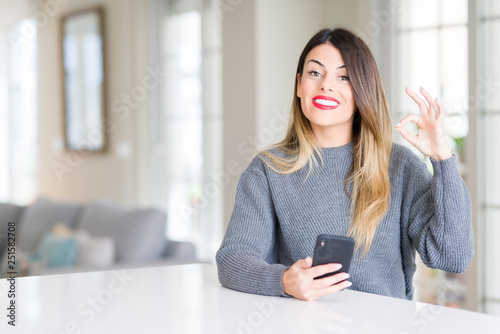Young beautiful woman using smartphone at home doing ok sign with fingers, excellent symbol