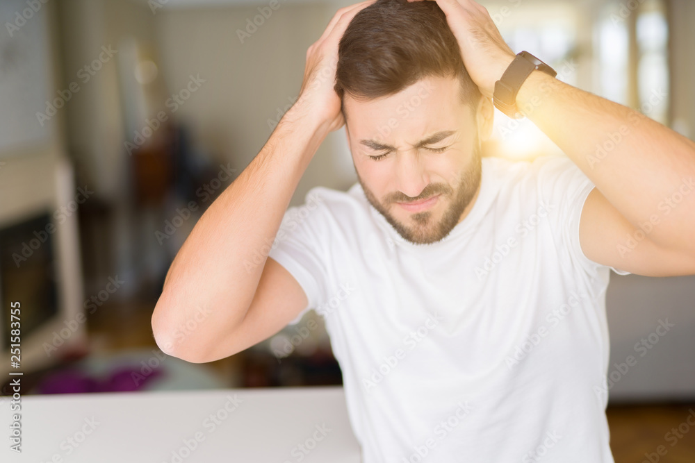 Young handsome man wearing casual white t-shirt at home suffering from headache desperate and stressed because pain and migraine. Hands on head. Young handsome man wearing casual white t-shirt at home suffering from headache desperate and stressed because pain and migraine. Hands on head.