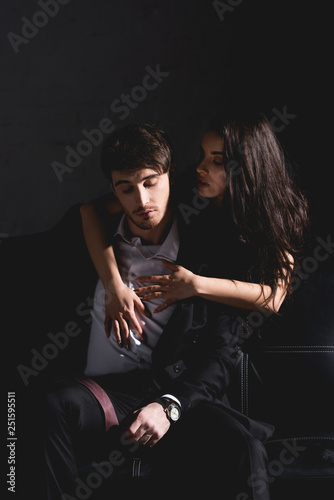brunette woman in black dress standing behind couch and hugging handsome man sitting on black background