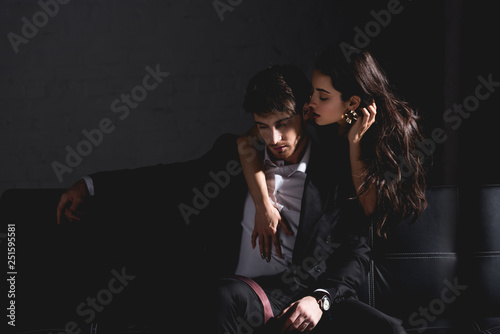 brunette woman in black dress standing behind couch, hugging and kissing handsome man in suit sitting on black background