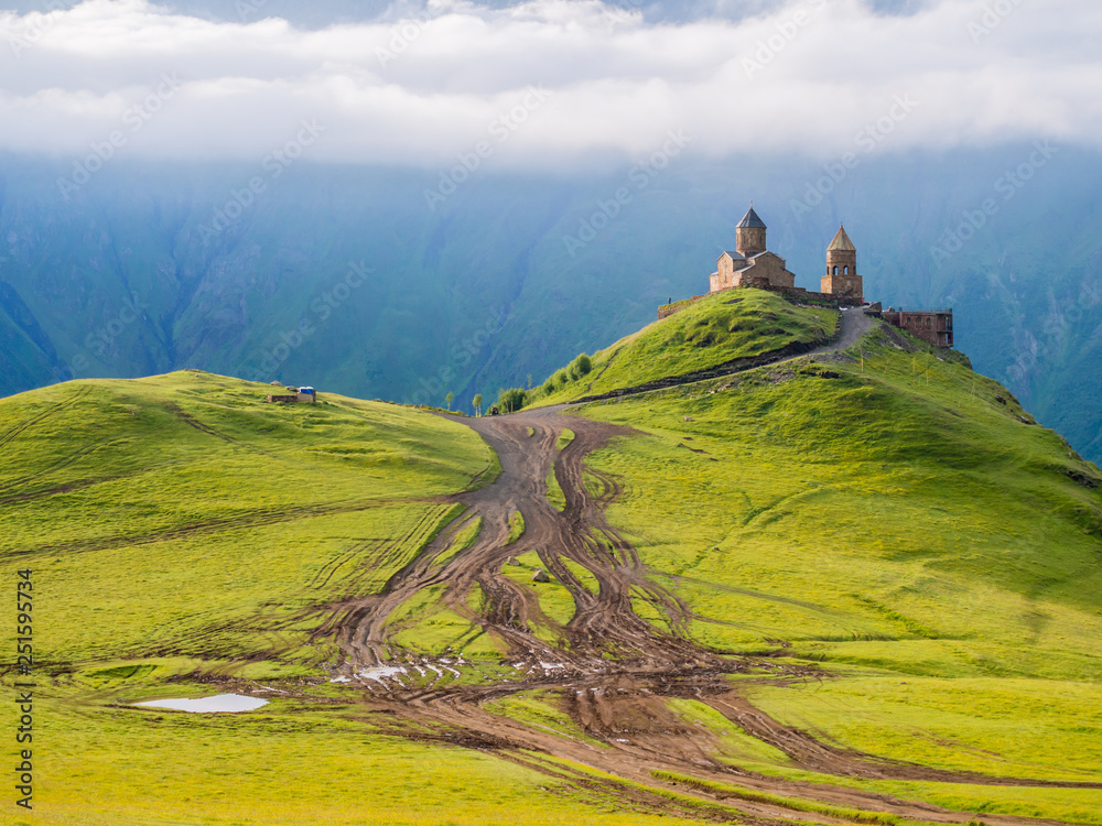 Colorful horizontal photo of Gergeti Tsminda Sameba church in Kazbegi (Stepancminda) village, Georgia, Caucasus, on a spring morning. Juicy green grass covers the hills, white clouds above the church.