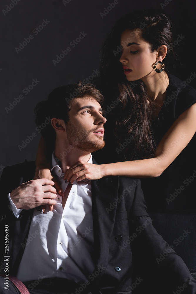 attractive woman in dress standing behind couch and hugging handsome man sitting on black background attractive woman in dress standing behind couch and hugging handsome man sitting on black background
