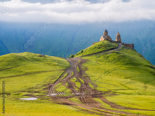 Obraz na płótnie Colorful horizontal photo of Gergeti Tsminda Sameba church in Kazbegi (Stepancminda) village, Georgia, Caucasus, on a spring morning. Juicy green grass covers the hills, white clouds above the church. w salonie