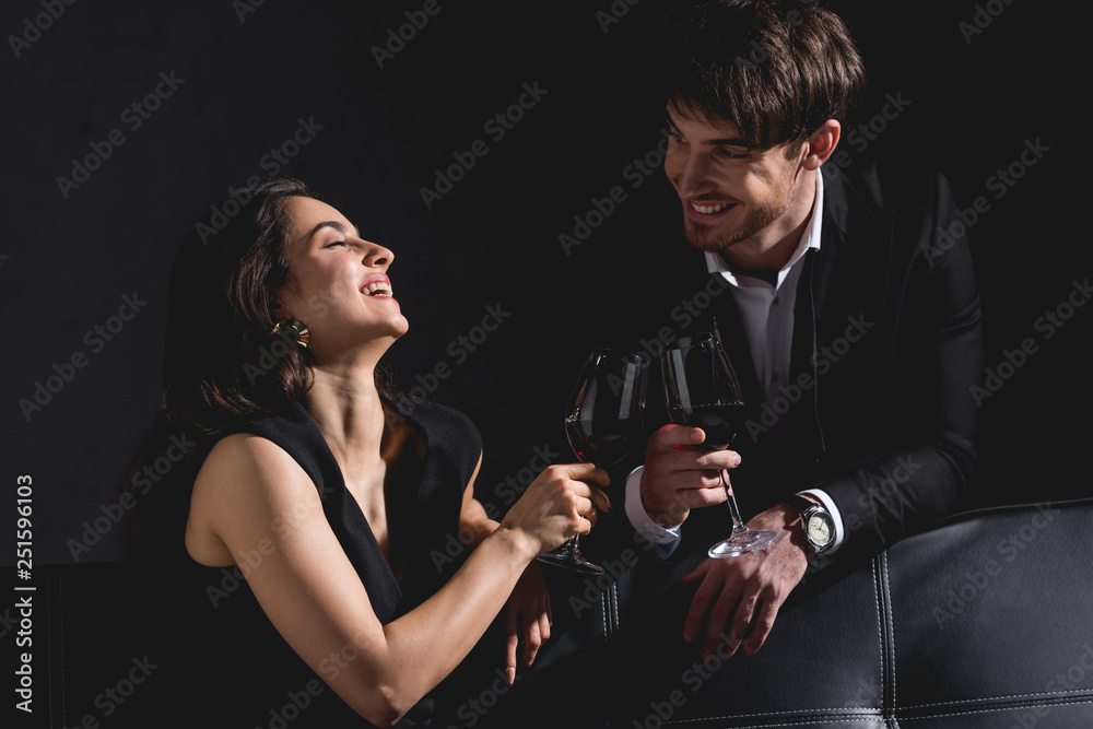 attractive woman sitting in dress, clinking glasses with handsome man standing behind couch and smiling on black background