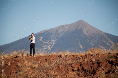 Young man in business attire holding a camera stands on a hill with a big mountain in the distance.