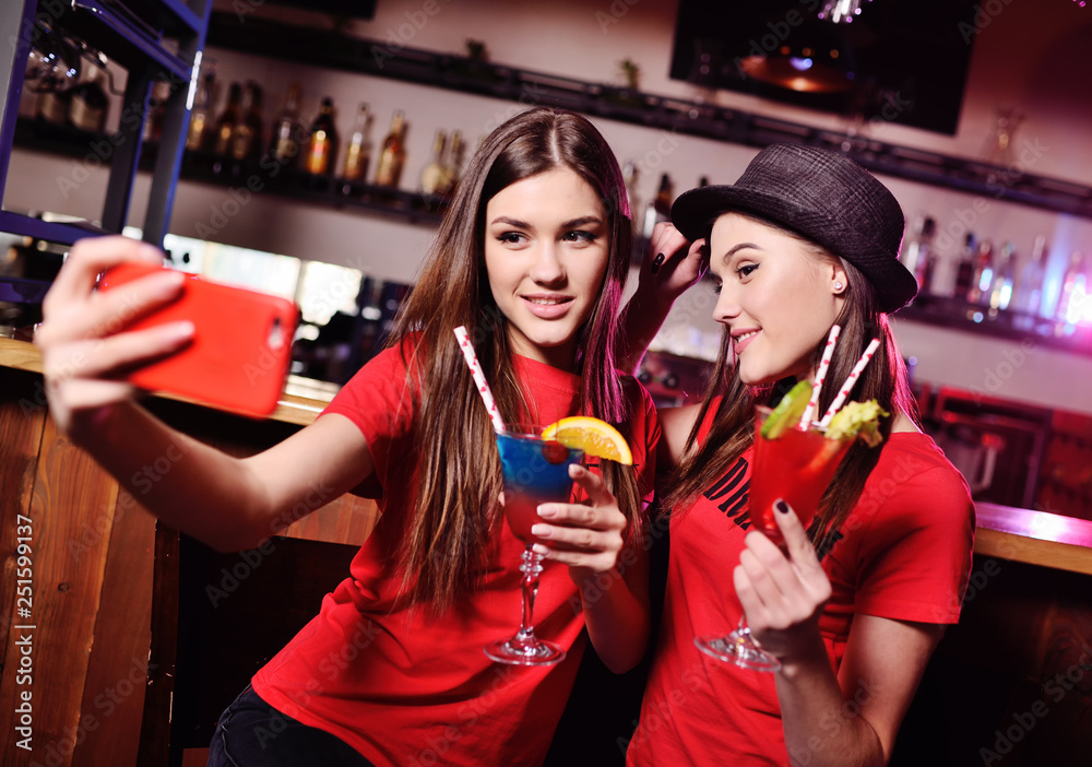 selfie time. two cute young girlfriends drink cocktails and are photographed on a smartphone camera on the background of a bar in a nightclub