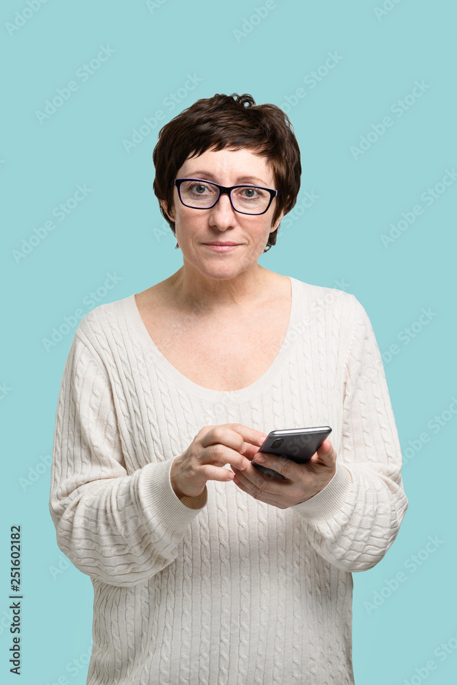 Adult woman in glass with smartphone in hand looking at camera and smiling on isolated blue background Adult woman in glass with smartphone in hand looking at camera and smiling on isolated blue background