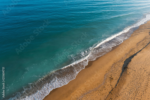 Scenic aerial view of golden sand waves and turquoise sea water with white foam from small waves. The concept of summer holidays. Bright abstract background perfect for any design. 