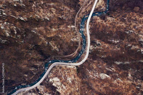 Aerial view of the mountain range, the flowing river and the paved highway. Winding road for cars among the mountains. Deciduous forest in the winter evening. Drone