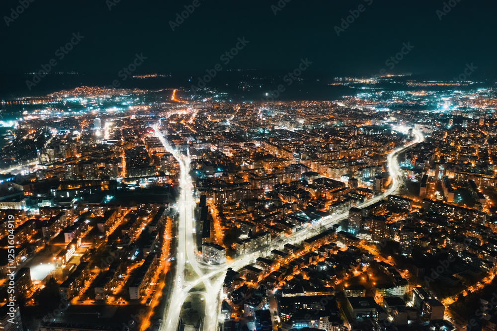 Aerial view of the resort town of Varna, Bulgaria at sunset of the day late in the evening. Bright lights of street lamps. Highways with car lights. Evening sky. The texture of the glowing night city