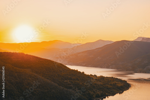 Panorama of the peaks of the beautiful tree-covered mountains at sunset on a sunny day in summer. Bright large orange sun disc. Solar reflection. A flash of the sun. Lens flare. Relax for tourist