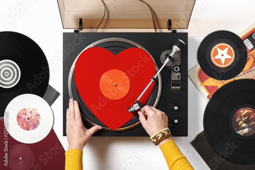 A red heart vinyl record turntable on a white table with plates. Included gramophone, torque. The hand of a girl DJ with bright accessory puts a stylus with a needle on a vinyl record. Valentine's Day photo