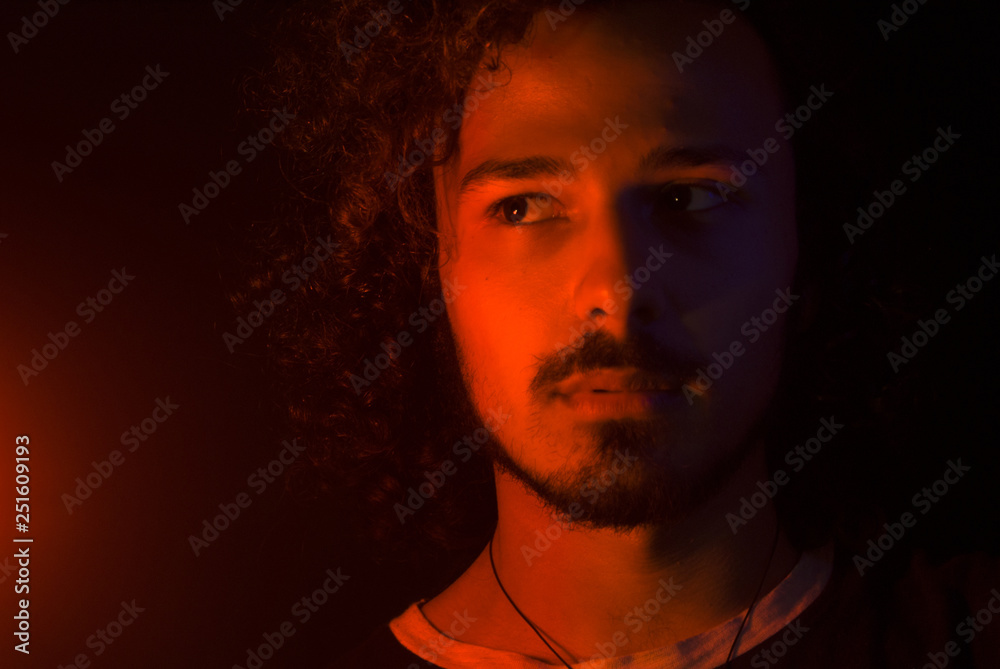 Portrait normal latin young man suspecting expression looking to the left with red light. Facial hair: moustache, beard, curly hair Portrait normal latin young man suspecting expression looking to the left with red light. Facial hair: moustache, beard, curly hair