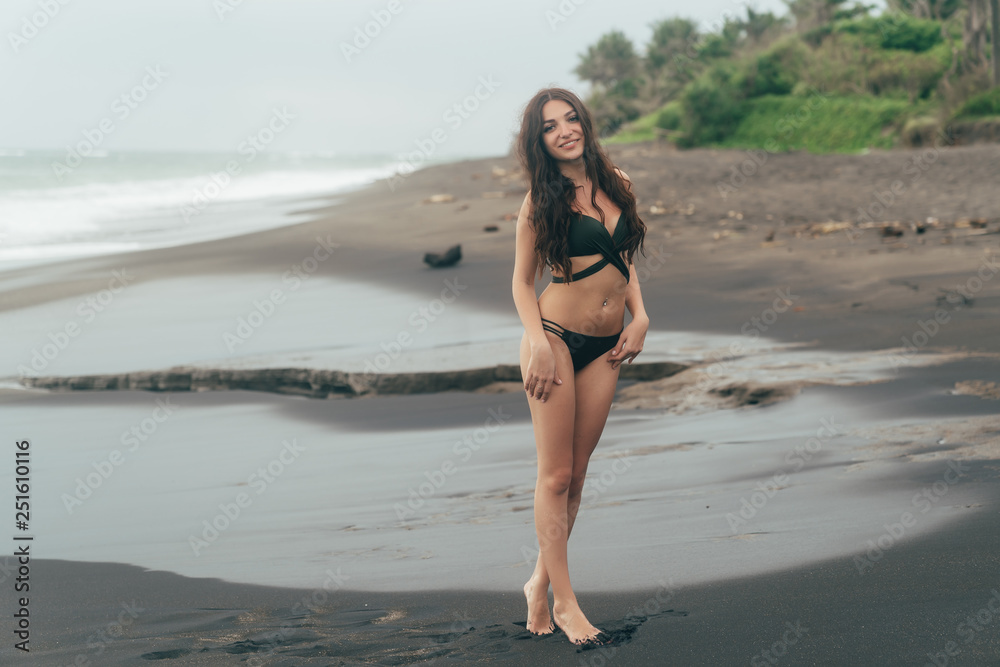 Sexy brunette girl in swimwear resting on black sand beach. Attractive model with gorgeous body posing near ocean