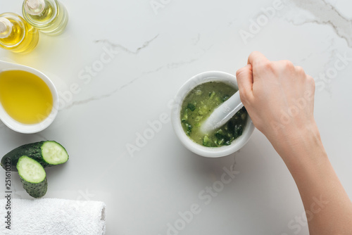 partial view of woman mixing cut cucumber with oil in pounder on white surface