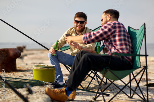 leisure and people concept - happy friends with smartphone and beer fishing on pier