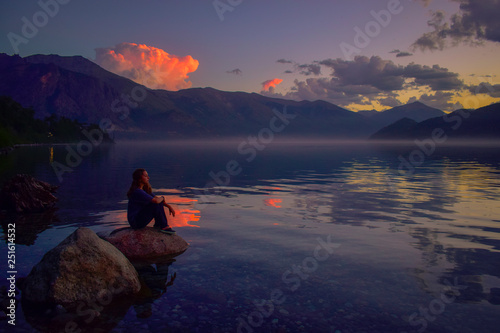 una chica a la orilla de un lago rodeado por montañas observa el atardecer