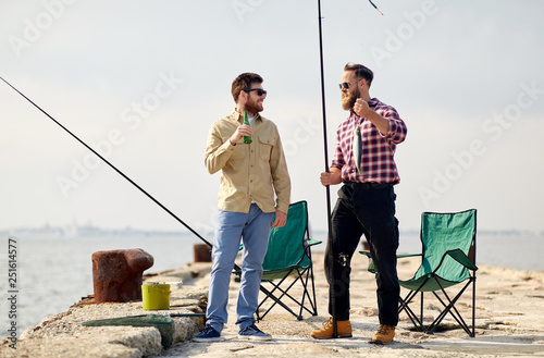 leisure and people concept - happy friends with fishing rods, fish and beer talking on pier at sea